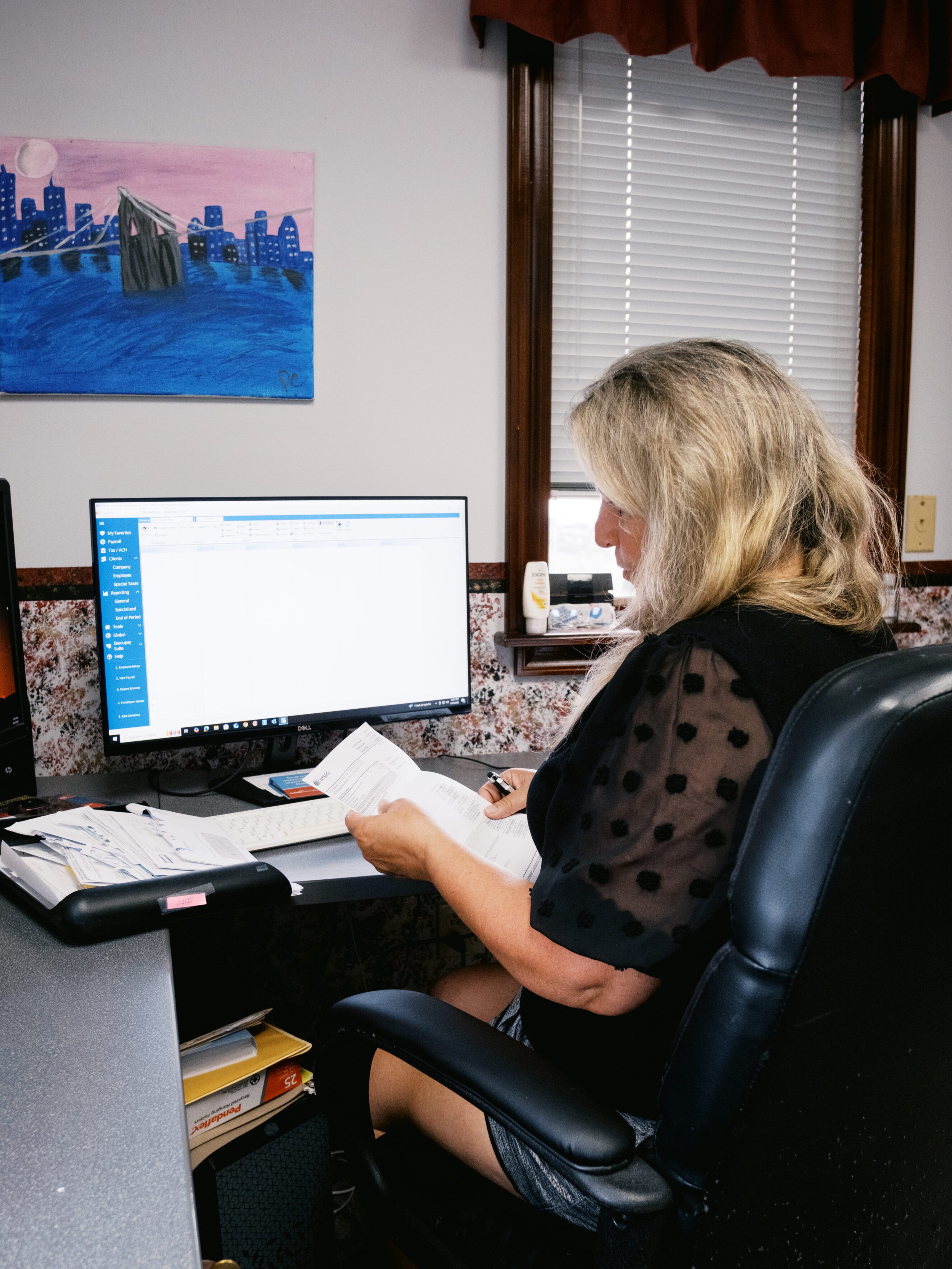 Woman reviewing payroll documents at her desk with a computer displaying payroll software, surrounded by office supplies and paperwork, illustrating efficient payroll management and compliance in a business setting.