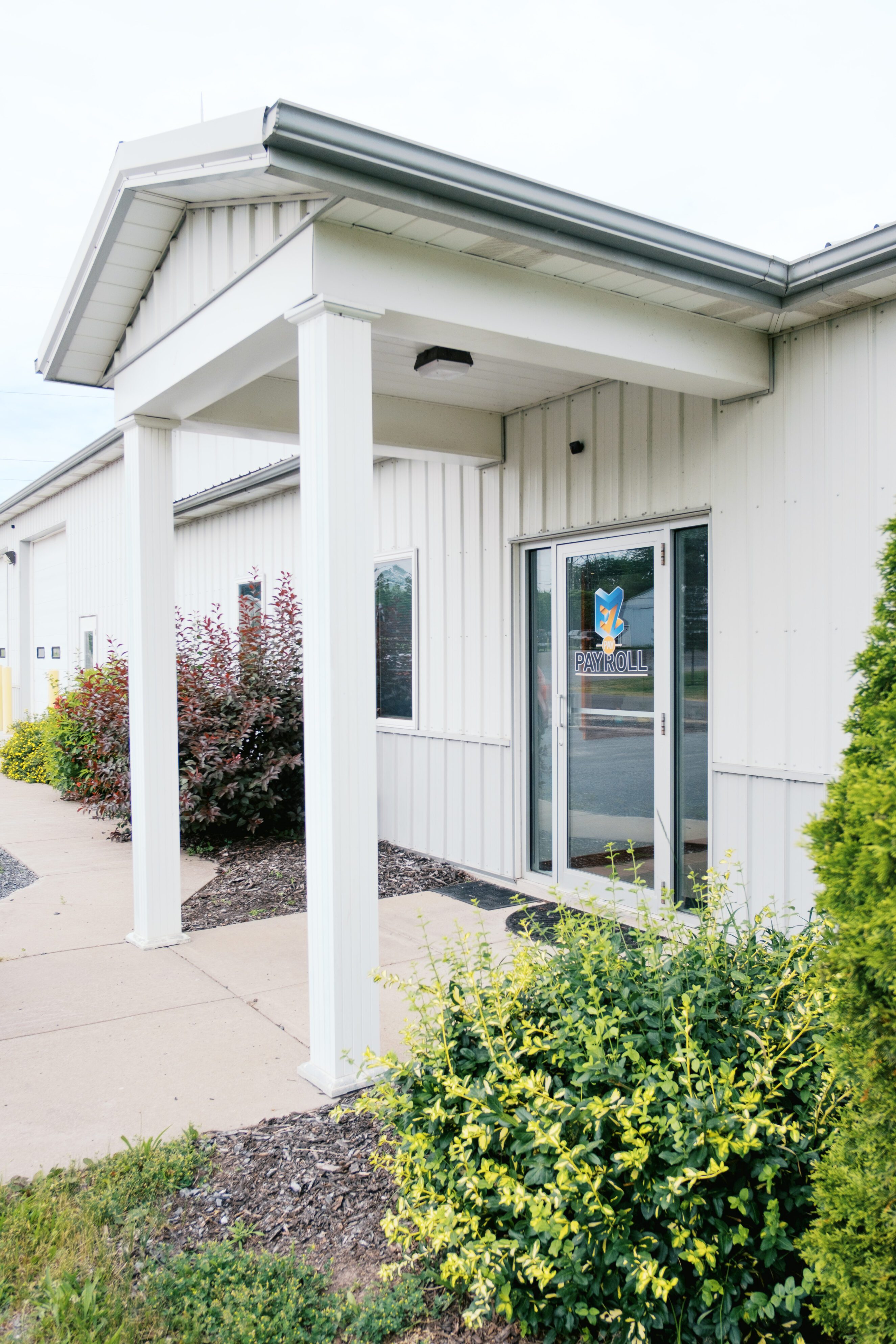 Entrance of EZPay Payroll office in Auburn, NY, showing white building exterior with glass door featuring company logo, surrounded by landscaped greenery and sidewalk.